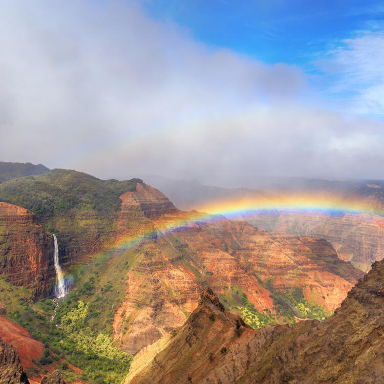 Atemberaubender Blick auf einen Regenbogen, der sich über den Waimea Canyon auf Kauai, Hawaii, spannt, mit markanten roten und grünen Schluchten