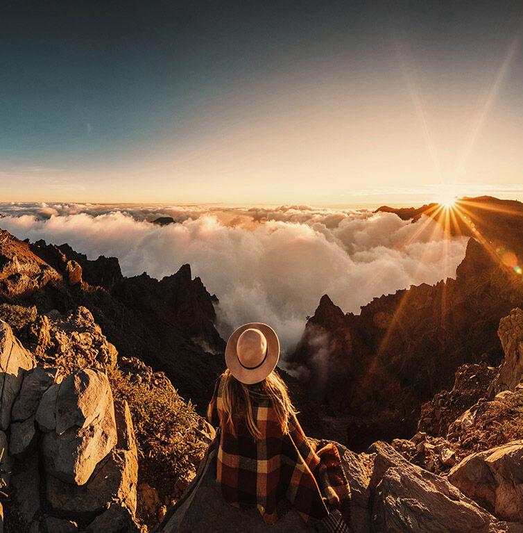 Eine Frau steht auf einem Berg, unter ihr die Wolkendecke, am Horizont geht die Sonne unter