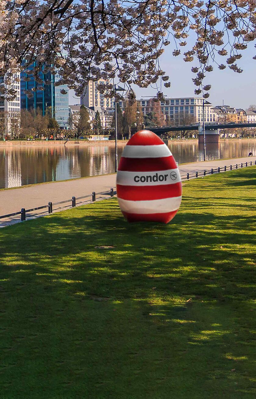 Huevo a rayas rojas y blancas y el logo de Condor sobre la hierba de un parque con el río y la ciudad de fondo