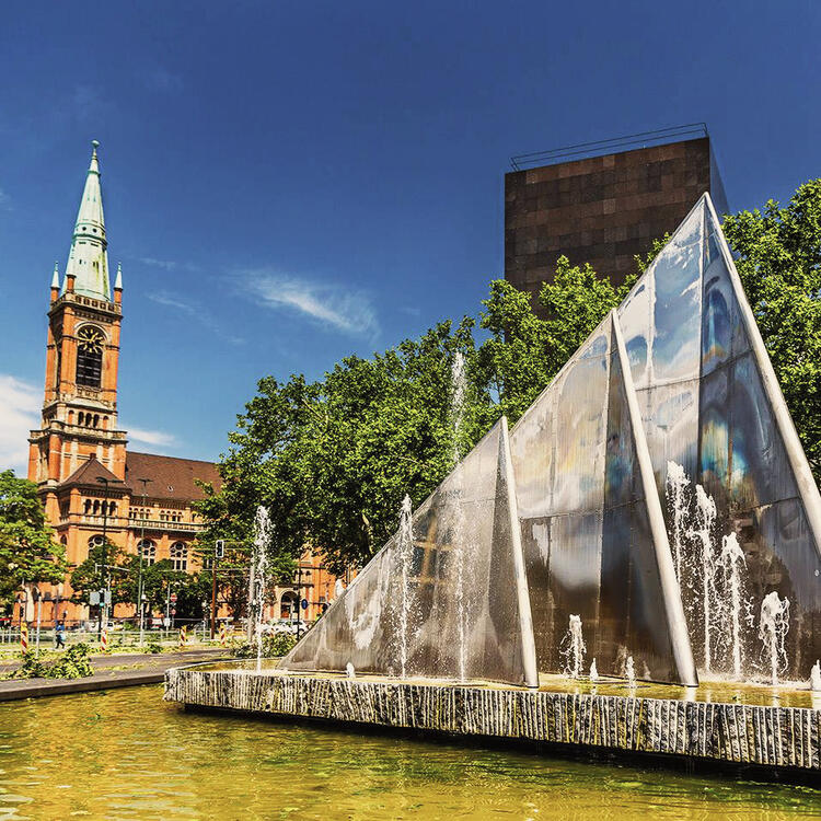 Fonte moderna em forma de pirâmide de vidro com jatos de água em primeiro plano, com a torre de uma igreja histórica e árvores verdes ao fundo sob um céu azul em Düsseldorf, Alemanha