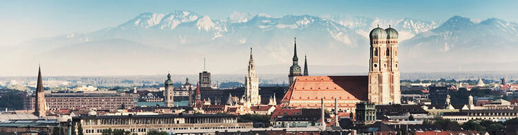 Panorama da cidade de Munique com os Alpes nevados ao fundo, destacando-se as torres gêmeas da Catedral de Frauenkirche e a arquitetura histórica variada sob um céu claro