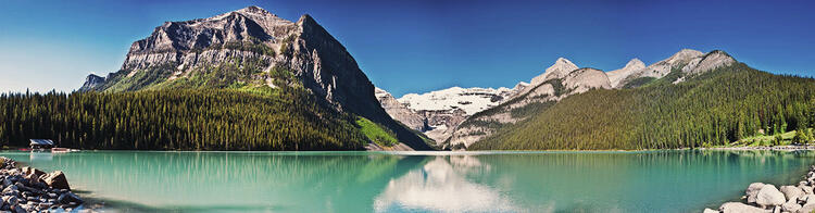 Paisagem panorâmica de um lago de águas turquesa com montanhas florestadas e um céu azul sem nuvens em um dia ensolarado