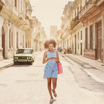 A woman walking down a vintage street, checking her phone, with classic architecture and an old car in the background