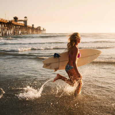 Female surfer running into the ocean at sunset near a pier with a surfboard in hand