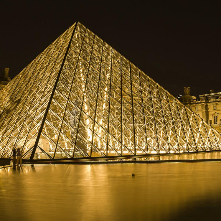  The Louvre Pyramid illuminated at night, reflecting off the water's surface with the Louvre Museum in the background.