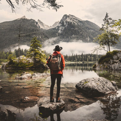 Traveler in a hat and backpack standing on a rock by a serene mountain lake with forest and misty peaks in the background