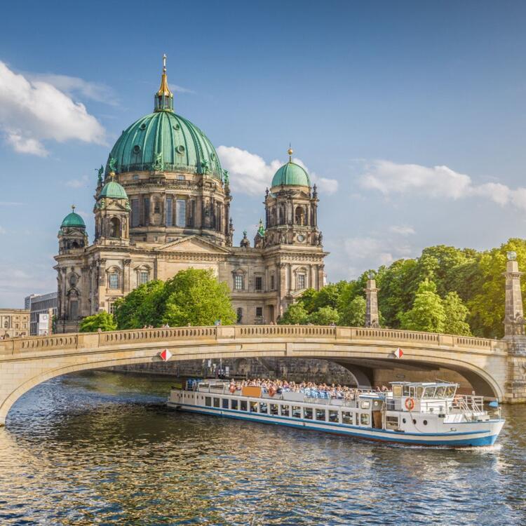 View of the buildings on the Spree Island with the Berlin Cathedral and its dome in the foreground and a passing tourist boat. 