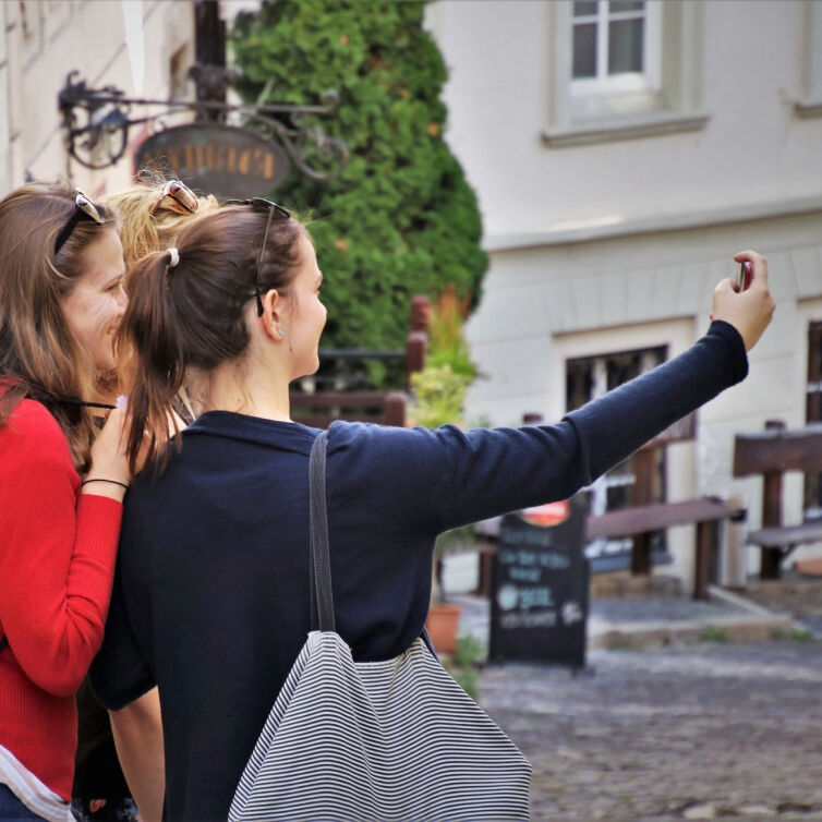 Two women taking a selfie together, one with a striped bag, capturing a moment of their travel adventures on a quaint city street