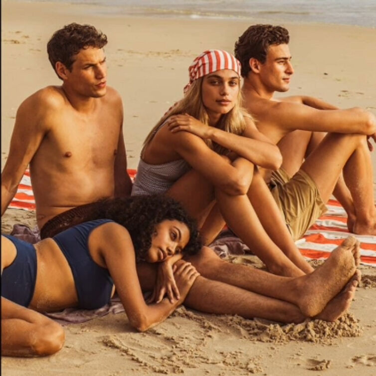 A group of young adults relaxing on a beach towel, with one woman lying in the sand, enjoying the company of friends on a sandy beach.