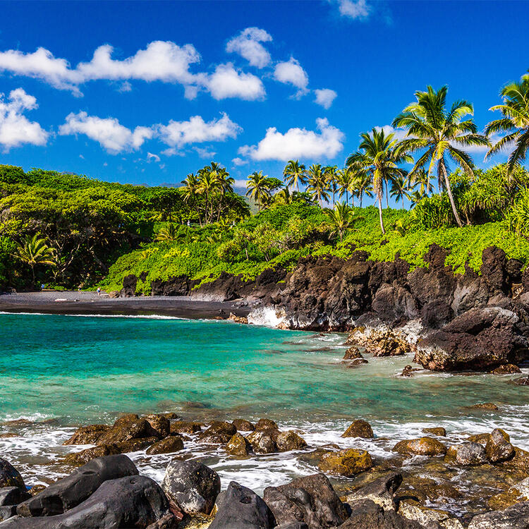 Waianapanapa State Park auf der Insel Maui in Hawaii - Im Vordergrund sind Felsen zu sehen, die sich in das türkisfarbene Wasser erstrecken, während im Hintergrund die Berge und die grüne Vegetation der Insel zu erkennen sind