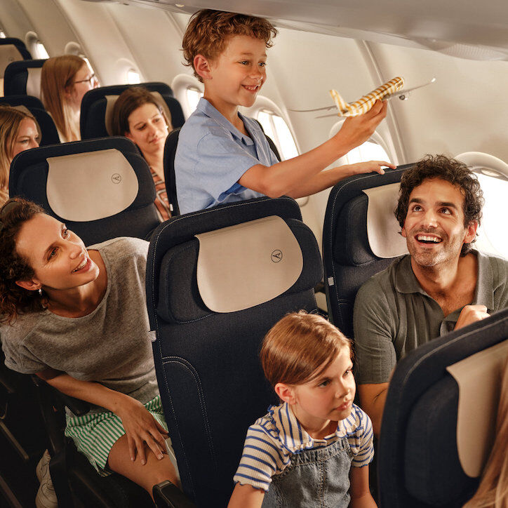 Ein junges Paar läuft die Sitzreihe in einem Condor-Flugzeug entlang; sie trägt ein rotes Sommerkleid und er einen Strohhut.