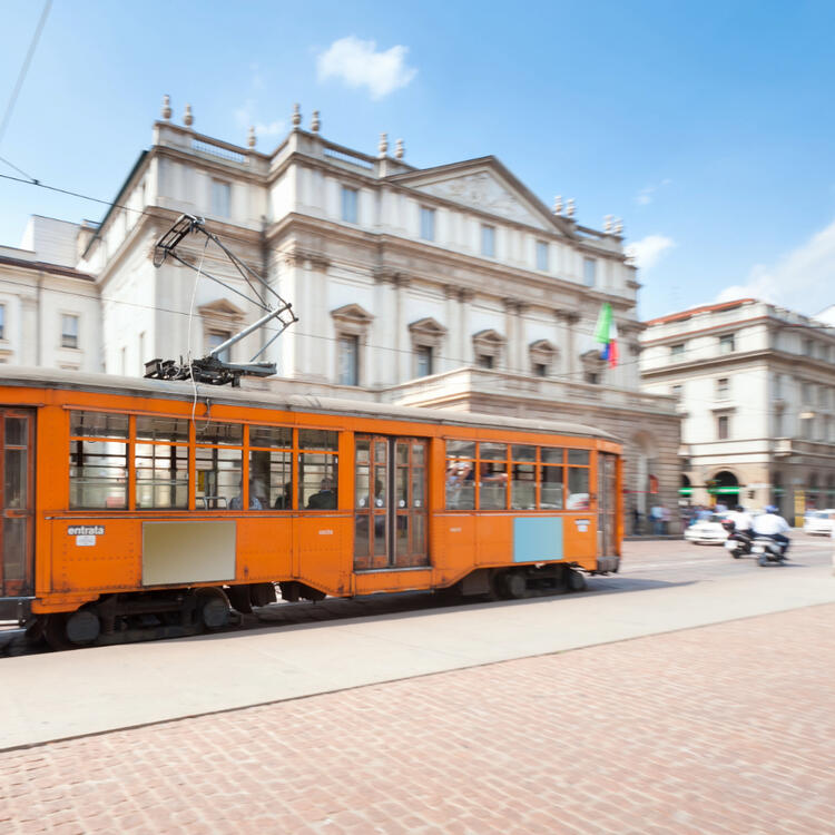 Ansicht des Theaters in Mailand mit einer orangefarbenen Straßenbahn im Vordergrund und blauem Himmel