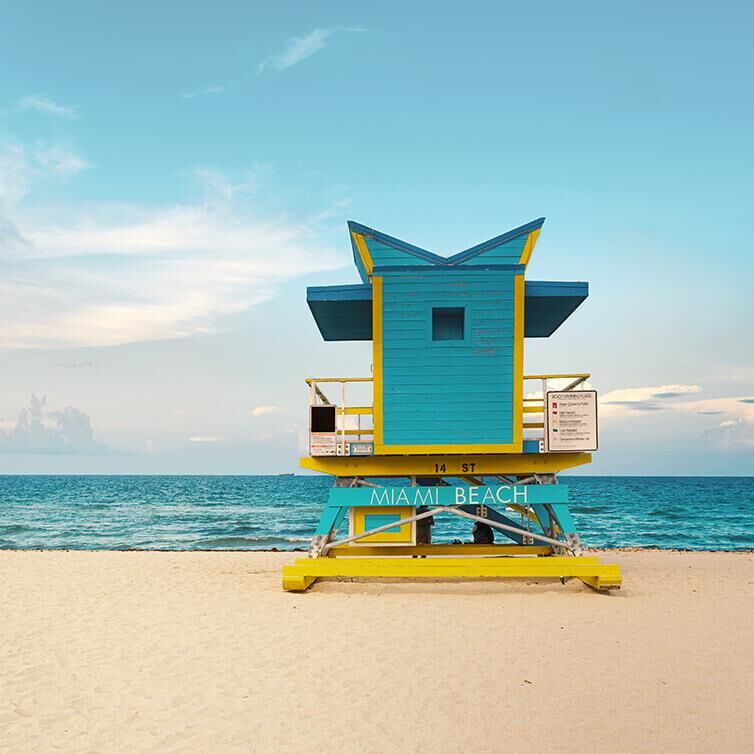 Ansicht vom Strand von Miami Beach, mit dem Meer im Hintergrund und dem goldenen Sand, mit einem blau-gelben Wachtposten und einem Schild, auf dem 'Miami Beach' steht.