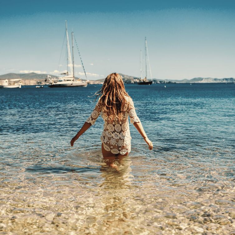 Eine Frau in einem weißen Hippie-Kleid steht im Meer auf Ibiza; sie breitet die Arme aus und schaut in die Sonne am blauen Himmel.