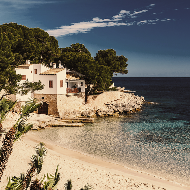 Eine Bucht auf Mallorca mit weißem Sand und türkisblauem Meer; über der Bucht ein typisches mallorcinisches Haus.