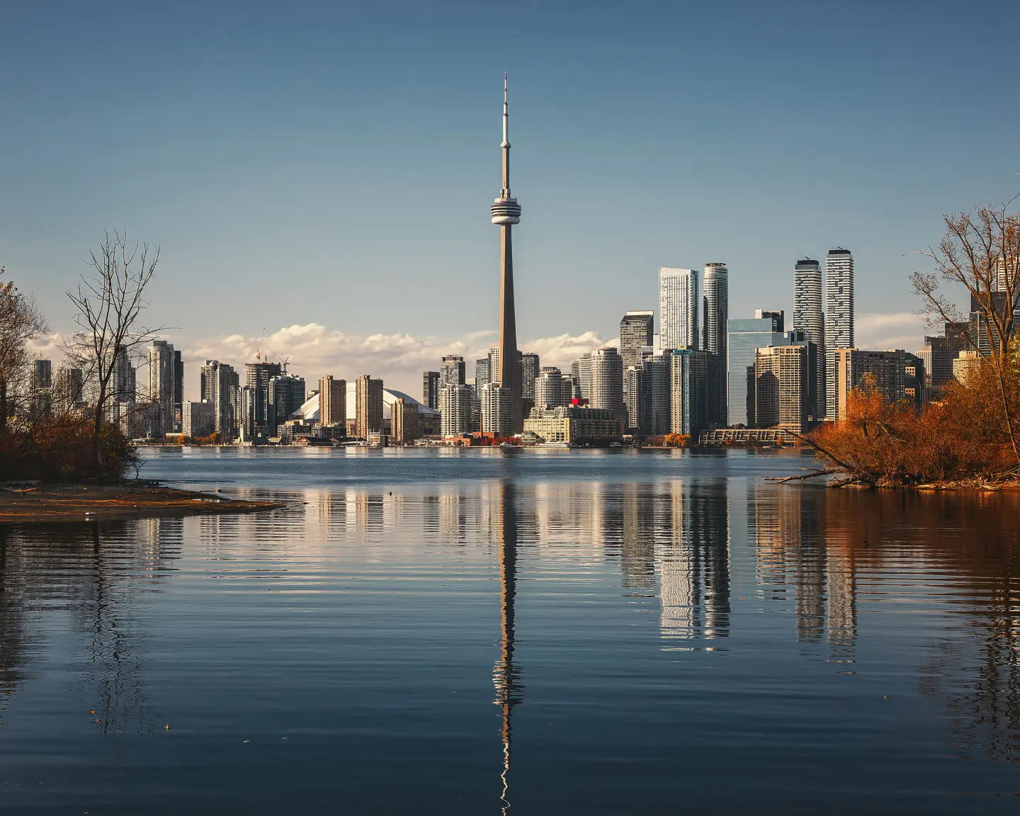 Panorama Toronta s věží CN Tower odrážející se ve vodách jezera Ontario obklopeného stromy v podzimních odstínech.