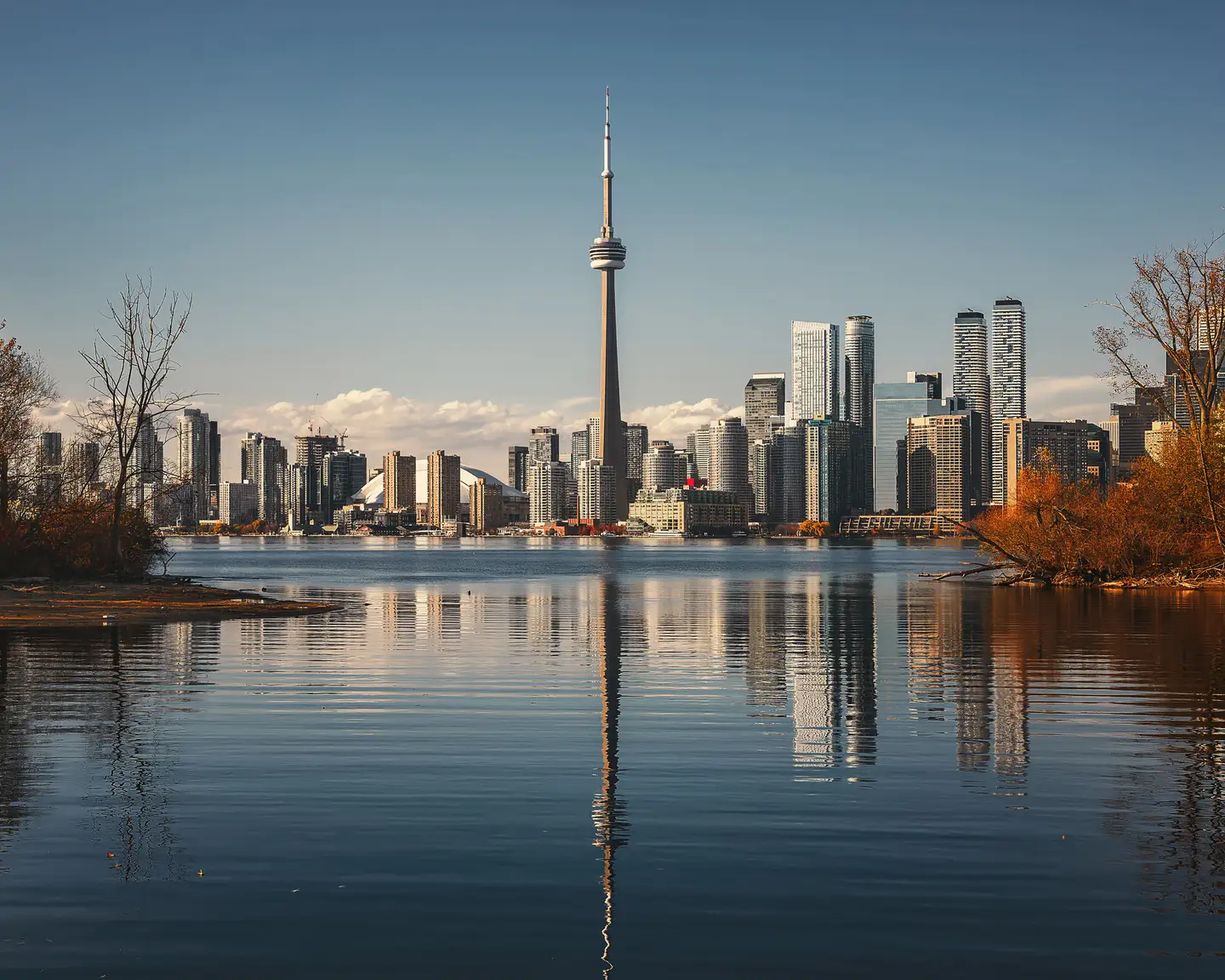 De skyline van Toronto met de CN Tower weerspiegeld in de wateren van Lake Ontario, omgeven door bomen in herfsttinten.