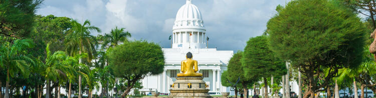 Statue dorée de Bouddha tournée vers l’hôtel de ville blanc de Colombo, entourée d’arbres tropicaux.