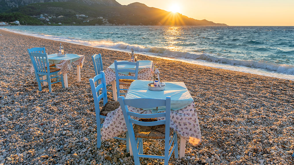 Zwei Tische mit Stühlen stehen am Kiesstrand von Kokkari, Abendstimmung