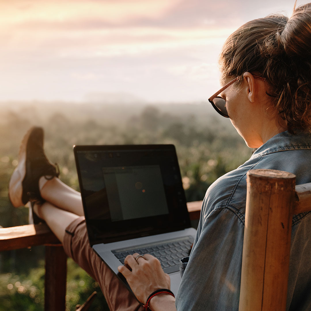 Frau mit Laptop auf den Beinen sitzt auf Balkon mit tropischer Landschaft im Hintergrund