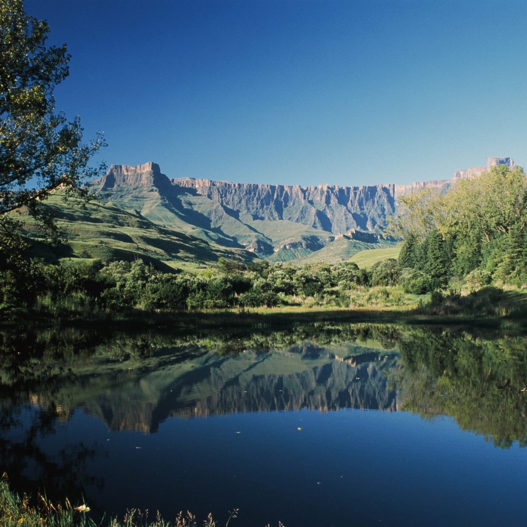 Spiegelglatter See vor grüner Berglandschaft und blauem Himmel.