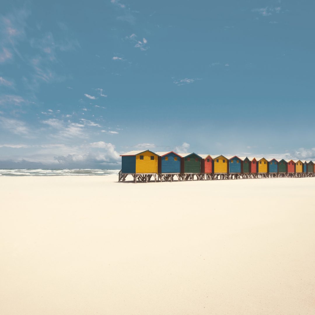 Bunte Strandhütten stehen in einer Reihe am weißen Sandstrand unter blauem Himmel.