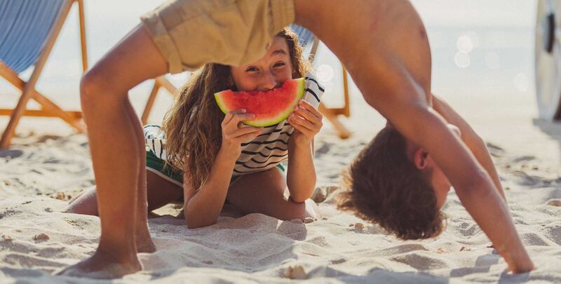 Ein Junge macht eine Brücke, ein Mädchen liegt darunter im Sand und beißt in ein Stück Wassermelone
