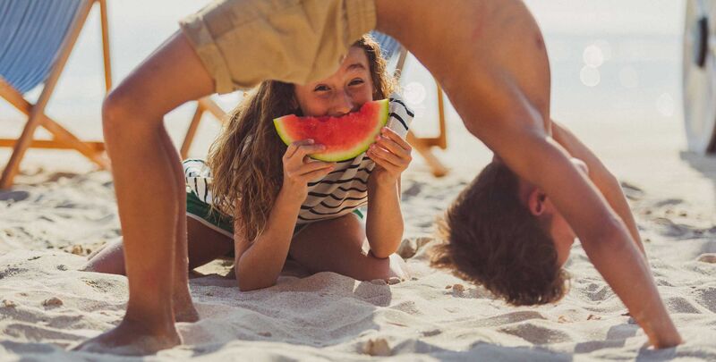 Strände in Griechenland Ein Junge macht eine Brücke, ein Mädchen liegt darunter im Sand und beißt in ein Stück Wassermelone