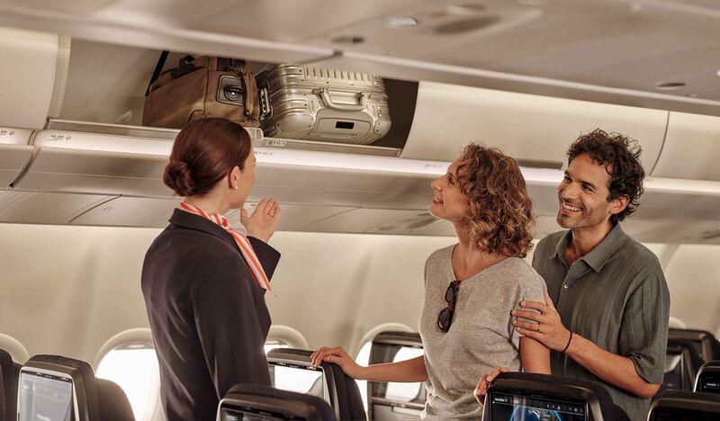 Eine Stewardess hilft zwei Urlaubern dabei, ihren Handgepäckskoffer im Gepäckfach über dem Sitz zu verstauen
