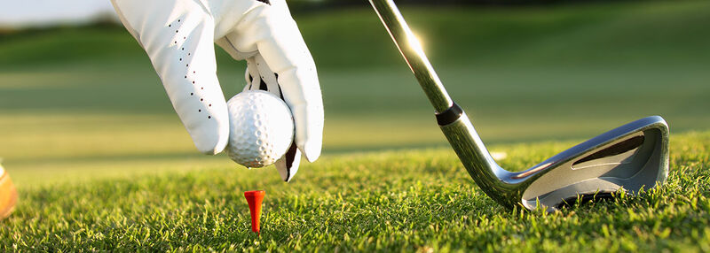 Eine Hand mit weißem Goldhandschuh setzt einen Golfball ein ein T. Daneben ist die Schlagfläche eines Schlägers zu sehen. 
