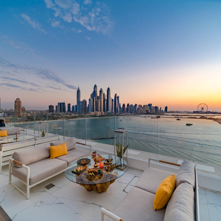 Aufnahme des Palm Jumeirah, umgeben von blauem Wasser, mit der Skyline der Stadt im Hintergrund unter einem klaren blauen Himmel.