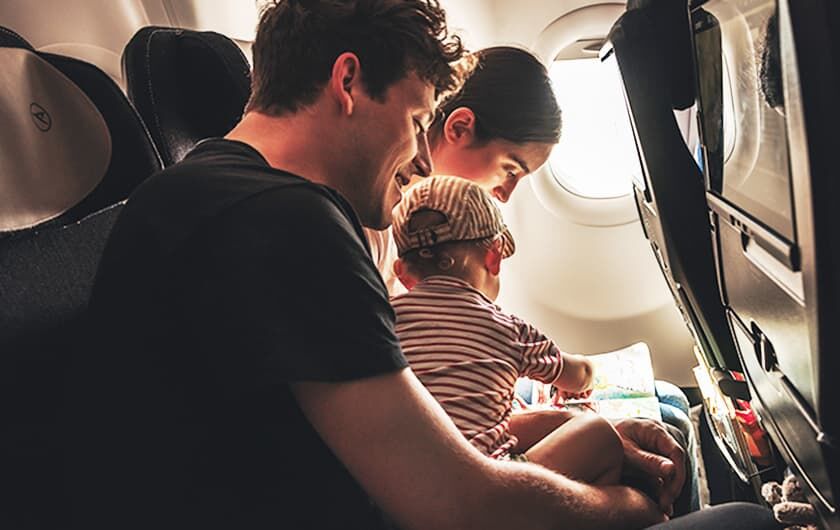 Eltern mit Kleinkind auf dem Schoß sitzen im Flugzeug und lesen ein Kinderbuch