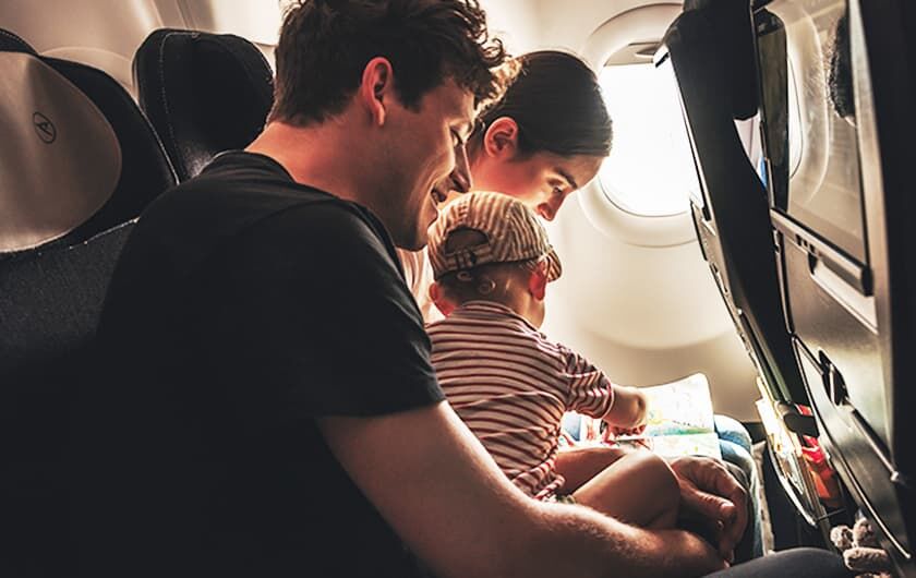 Fliegen mit Kleinkindern unter 2 Jahren Eltern mit Kleinkind auf dem Schoß sitzen im Flugzeug und lesen ein Kinderbuch