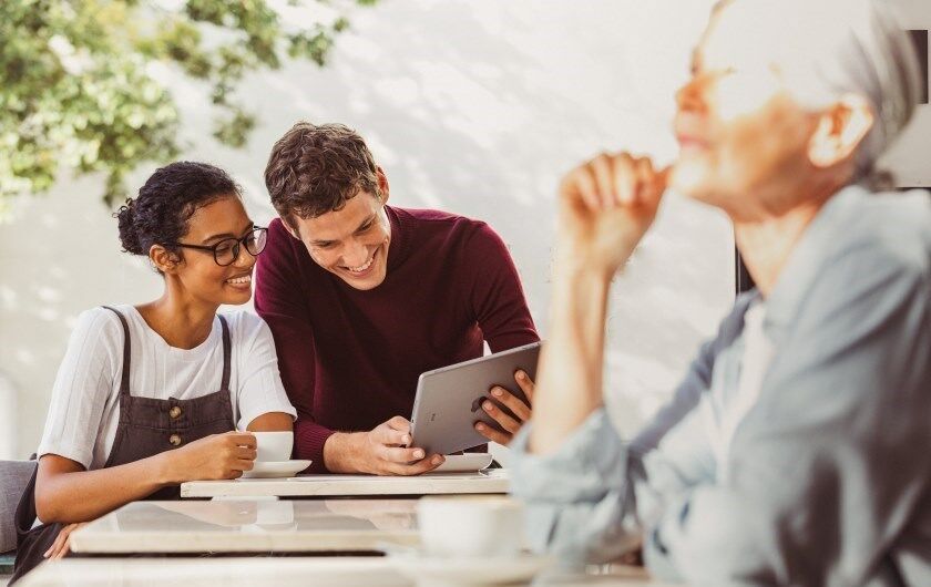 Young Talents | Praktikum oder Werkstudentenjob Ein Päarchen sitzt im Café. Sie schauen auf ein Tablet. Eine ältere DAme sitzt rechts weiter vorne allein an einem Tisch.
