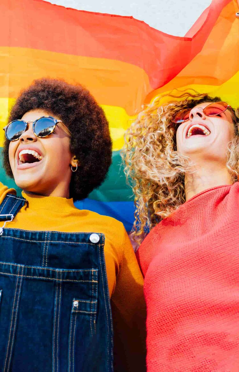 Zwei Frauen, die eine Regenbogenflagge in die Luft halten und lachen