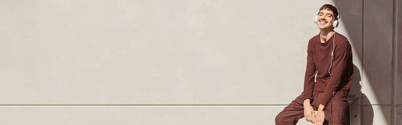 Ein Passagier sitzt glücklich auf seinem Koffer, er trägt große Kopfhörer und blickt mit geschlossenen Augen in die Sonne