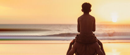 Ein Mann hat seinen Sohn auf den Schultern und schaut mit ihm aufs Meer hinaus, wo die Sonne gerade untergeht