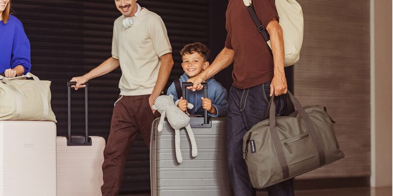 Condor Priority Package Zwei Männer, eine Frau und ein Kind, das einen Stofftier-Affen im Arm hält, sind lachend mit ihren Koffern unterwegs.
