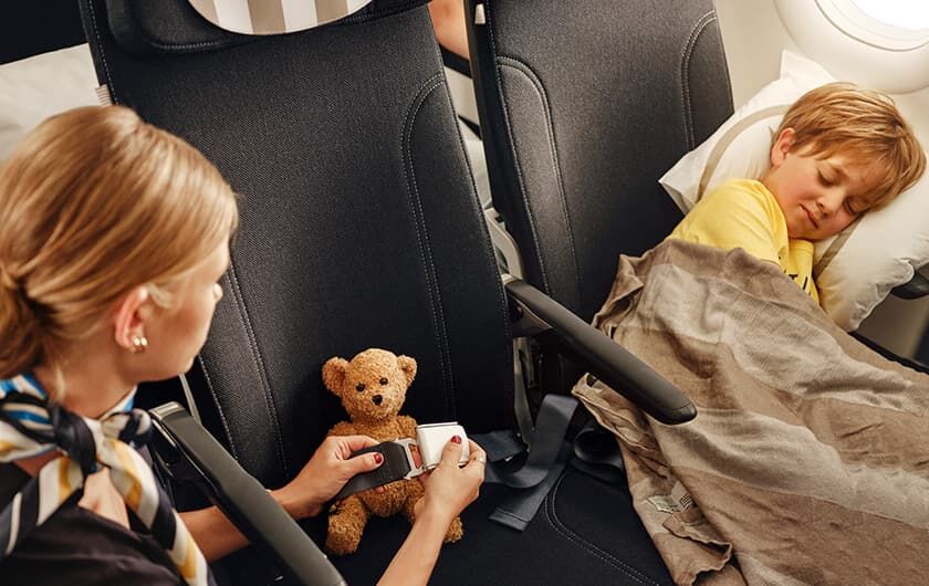 Stewardess schnallt kleinen Bären im Sitz des Flugzeuges fest, kleiner Junge liegt auf dem Nebensitz und schaut zu