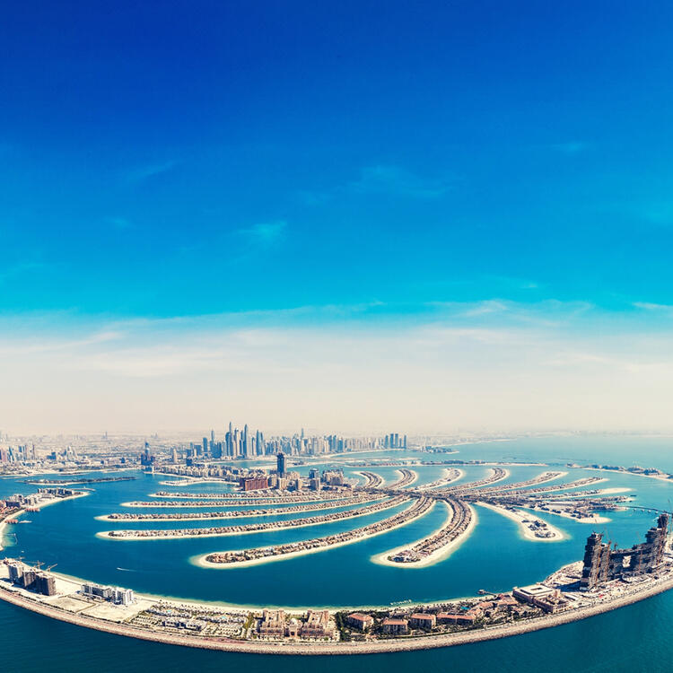 Luftaufnahme von Palm Jumeirah in Dubai mit seiner charakteristischen Palmenform, umgeben von blauem Wasser, mit der Skyline der Stadt im Hintergrund unter einem klaren blauen Himmel.