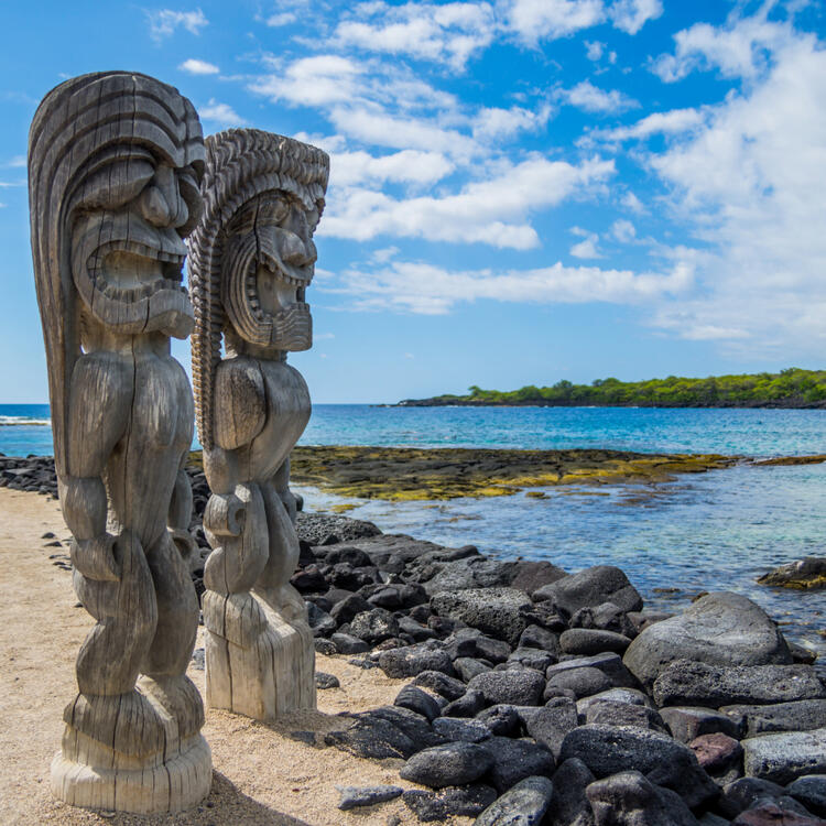 raditionelle hawaiianische Tiki-Statuen am Puʻuhonua o Hōnaunau National Historical Park mit Blick auf das türkisblaue Meer.