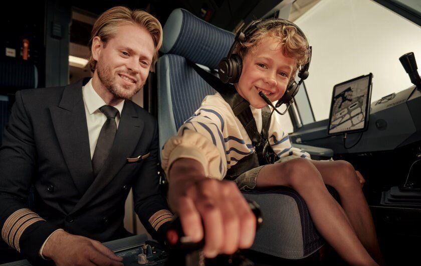 Kleiner Junge sitzt auf dem Platz des Piloten im Cockpit. Der Pilot kniet links neben dem Jungen. 