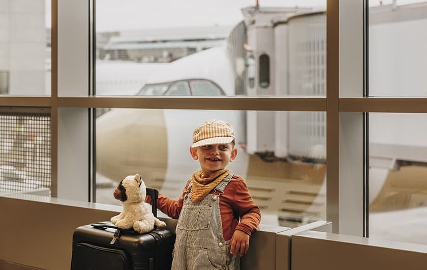 Kleinkind mit Koffer und Kuscheltier steht am Fenster des Flughafens, im Hintergrund ist ein stehendes Flugzeug