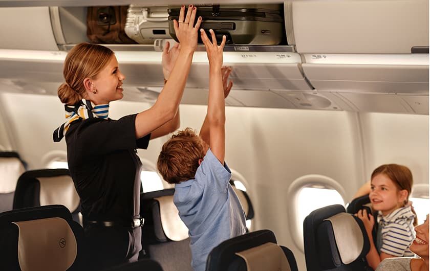 Stellenangebote Kabine Flugbegleiterin packt Koffer in die Gepäckablage. Ei Kind hilft ihr dabei.