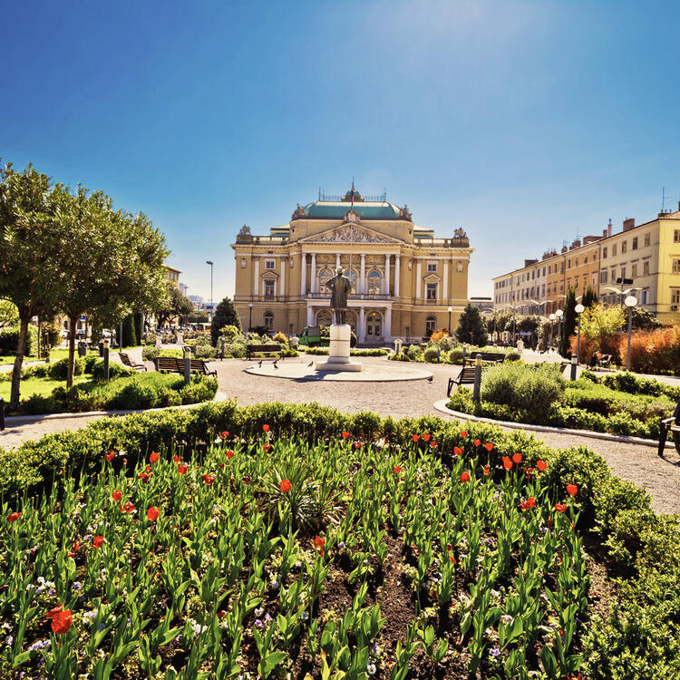 Großes Opernhaus mit neoklassischer Architektur, prominent platzierte Statue und üppige Gärten auf einem Stadtplatz