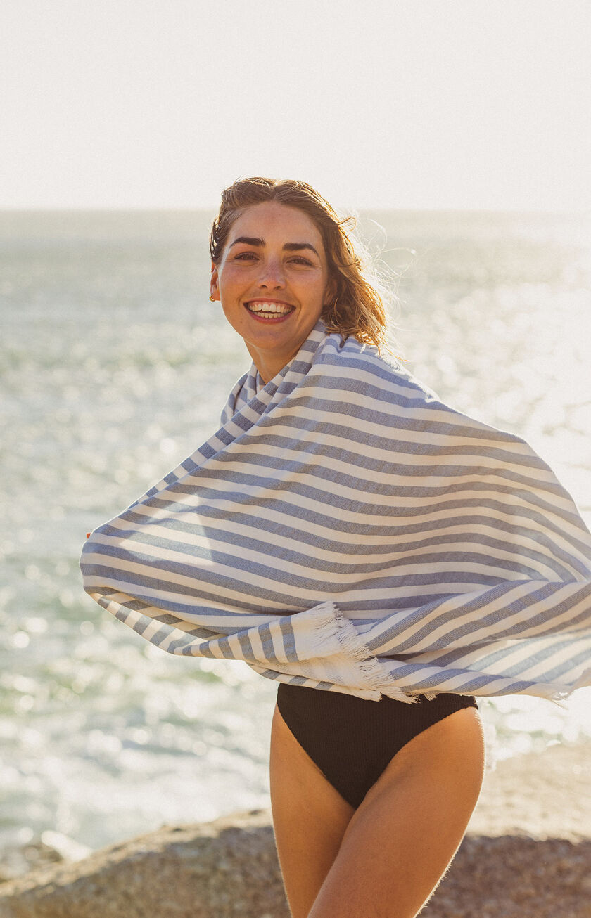 Eine Frau mit einem gestreiften Tuch um die Schultern am Strand, hinter ihr das von der Sonne glitzernde Meer