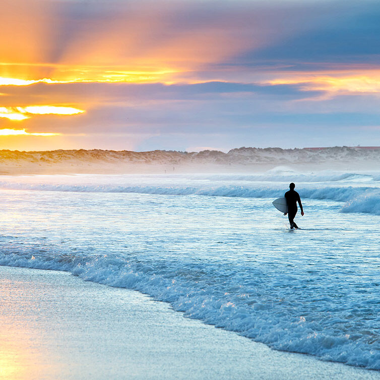 Persona con una tabla de surf saliendo del mar en una playa en Nazaré, Portugal, famosa por sus olas