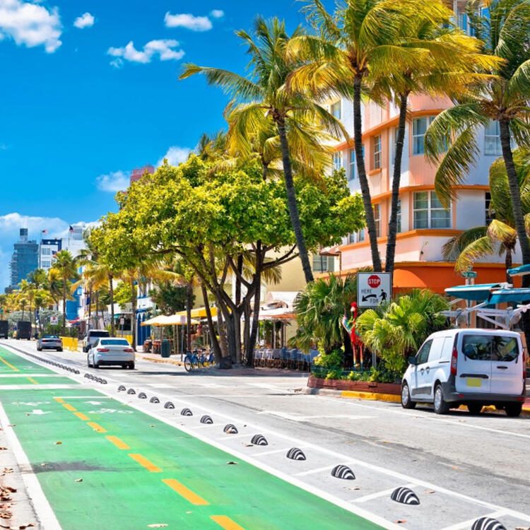 Una vibrante calle de Miami con un carril bici verde, palmeras y edificios de colores pastel bajo un cielo azul 