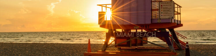 El cálido resplandor del amanecer ilumina una caseta de socorrista de Miami Beach, con los rayos del sol atravesando la estructura sobre un fondo de océano y cielo nublado.