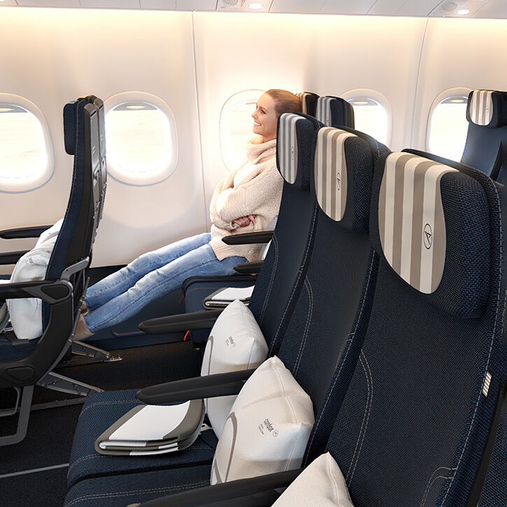 La foto muestra a una mujer en un vuelo de Condor en Clase Premium Economy, con las piernas estiradas y en una actitud relajada, sentada junto a la ventana. Los asientos tienen un reposacabezas y una manta, ambos a rayas en colores blanco y arena, y cuentan con almohadas blancas con el logo de Condor